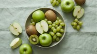 Fresh green fruits arranged in a ceramic bowl and on linen fabric, featuring apples, kiwi, and grapes in a natural flat lay composition.