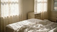 Cozy unmade bed with rumpled white bedding in warm sunlit bedroom with soft natural lighting streaming through lace curtains creating peaceful morning atmosphere.