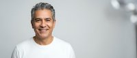 Confident mature Hispanic businessman in white shirt smiling warmly against modern office background for corporate marketing and professional content.