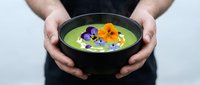 Hands holding black bowl with vibrant green soup garnished with edible flowers, culinary presentation for fine dining, restaurant menu, cooking blogs, food styling, and gourmet cuisine.