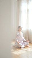 Young Asian woman in white pajamas meditating in lotus position on wooden floor by bright window with natural sunlight streaming through curtains.