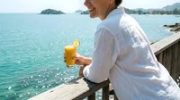 Man in white shirt holding fresh orange juice on wooden dock overlooking turquoise tropical water and mountains, summer vacation and relaxation concept.