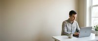 Young professional man working on laptop at modern desk in bright office space with natural light from window creating productive workspace atmosphere.