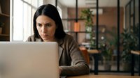 Focused businesswoman working on laptop in modern office space with natural lighting and industrial design elements for professional productivity.