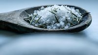 Coarse sea salt crystals with dried herbs on rustic wooden spoon for cooking seasoning and culinary preparation on neutral background.