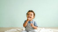 Happy toddler boy with curly blonde hair, approximately 18-24 months old, wearing gray apron over blue shirt, smiling and gesturing with hands while playing with flour on wooden surface against mint green wall.