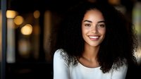 Young African American woman with natural curly hair and bright smile wearing white sweater against blurred bokeh background, radiating confidence and joy.