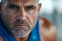 Close up portrait of mature athletic Caucasian male with gray beard and intense blue eyes wearing blue tank top, looking directly at camera with determined expression.