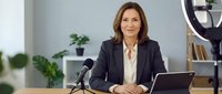 Middle-aged woman with brown hair and light skin, wearing dark blazer and cream blouse, seated at desk with microphone and laptop for podcast, interview, or broadcast recording in modern office setting.