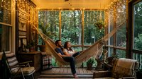 Young couple relaxing in hammock on wooden cabin porch with large windows overlooking forest, cozy interior design with warm lighting and natural surroundings.