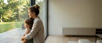 Caucasian mother gently hugging her young son while both gaze through large windows at a lush green garden in a modern minimalist home.