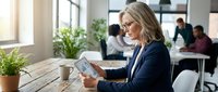 Professional blonde businesswoman using smartphone in modern office workspace with colleagues working in background.