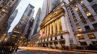 Iconic Wall Street financial district with illuminated New York Stock Exchange building facade at dusk, light trails from passing traffic create dynamic movement.