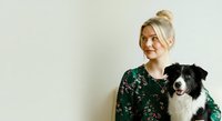 Young woman with blonde hair in a bun, fair complexion, wearing a dark green floral blouse, posing with a black and white border collie dog against a neutral background for pet care and lifestyle content.