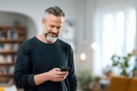 Mature Caucasian man with gray beard smiling while using smartphone in modern home interior. Happy middle-aged male texting on mobile phone indoors.