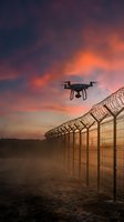 Drone flying over security fence with barbed wire at dramatic sunset for surveillance and border protection technology applications.