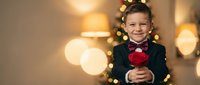 Young boy in formal suit with bow tie holding red rose near Christmas tree with warm bokeh lights creating festive holiday atmosphere for greeting cards.