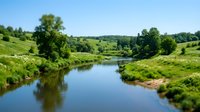 Scenic river flowing through lush green countryside landscape with trees and rolling hills under clear blue sky, suitable for nature and travel content.