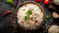Steamed basmati rice with red chili peppers and fresh herbs in wooden bowl on dark background, surrounded by cooking ingredients.