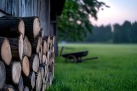 Stacked firewood logs against rustic barn wall with blurred wooden bench on misty green meadow at dawn, creating peaceful countryside atmosphere.