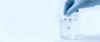 Hand in blue latex glove dropping white tablets into glass beaker with water on light background, pharmaceutical research concept.