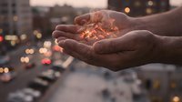Cupped hands holding glowing light particles against blurred city traffic at dusk, symbolizing hope, protection and magical energy in urban environment.