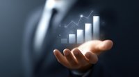 Business professional's hand holding glowing digital graph showing upward trend with rising bar chart and line graph floating above palm against dark background.