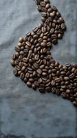 Roasted coffee beans scattered on gray linen fabric creating organic pattern for cafe branding and beverage marketing materials.