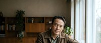 Mature Asian man in brown shirt sitting thoughtfully by window in modern home office with wooden shelves and plants creating warm professional atmosphere.