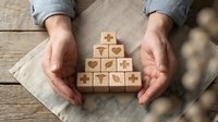 Hands protecting wooden blocks with healthcare medical symbols arranged in pyramid shape on rustic wood background for wellness insurance concept.