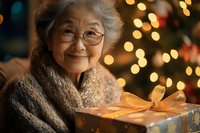 Smiling senior Asian woman holding golden wrapped Christmas present against festive bokeh lights background, wearing cozy knit scarf and glasses.