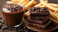 Rich chocolate mousse in glass with dark chocolate chunks on wooden plate and french fries in background, selective focus on dessert with soft blur.