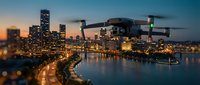 Modern drone flying over illuminated city skyline at twilight with river reflections and urban lights creating atmospheric evening scene.