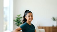 Young woman with dark hair pulled back, South Asian ethnicity, wearing dark teal t-shirt, smiling at camera in bright modern home interior with plants and natural light.