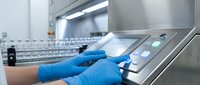 Technician in blue gloves operating laboratory analyzer machine in sterile medical facility, conducting diagnostic testing and sample analysis for healthcare research.