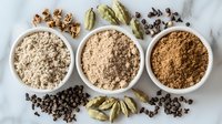 Ground spices and whole ingredients arranged in white bowls on marble surface, featuring cardamom pods, black peppercorns and aromatic powder mixtures.