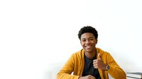 Young man with dark curly hair and medium brown skin tone wearing a mustard yellow jacket over a black shirt, giving thumbs up gesture against white background for approval and success concepts.