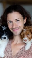 Caucasian male with long dark hair and blue eyes smiling while holding two adorable puppies with matching blue eyes, expressing joy and companionship in soft lighting.