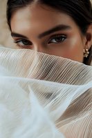 Middle Eastern woman with expressive brown eyes and defined eyebrows partially covered by sheer white pleated fabric, creating mysterious and elegant portrait.