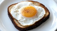 Sunny side up egg on dark toasted bread slice seasoned with black pepper and salt, served on white plate. Close up view shows runny yolk and crispy edges.