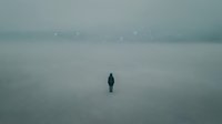 Solitary figure standing alone in vast misty landscape with ethereal light particles floating through fog, conveying isolation and contemplation for conceptual and editorial use.