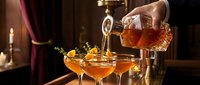 Bartender pouring amber cocktail from crystal decanter into coupe glasses with orange garnish in upscale bar setting with warm atmospheric lighting.