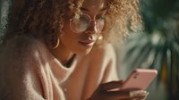 Young woman with curly hair and glasses using smartphone in soft evening light with tropical plants in background.