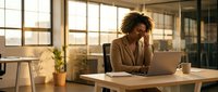 Young African American businesswoman working on laptop in modern office space with natural lighting and glass walls for corporate presentations.