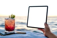 Person holding blank tablet screen at beach sunset with red cocktail garnished with lime on sand, mockup for digital display presentation and summer lifestyle.