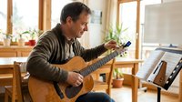 Middle-aged Caucasian man playing acoustic guitar at home, reading sheet music on stand. Cozy living room with natural light from windows. Music hobby concept.