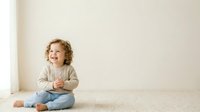 Toddler with curly blonde hair, Caucasian, wearing beige cardigan and jeans, sitting on carpet smiling. Suitable for parenting, childcare, and family lifestyle content.