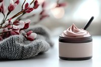 Pink cosmetic cream in black jar with knitted wool scarf and spring pink flowers on blurred background, luxury skincare product photography.