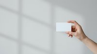 Female hand holding blank white business card against neutral wall with window light shadows, suitable for branding and corporate identity mockups.