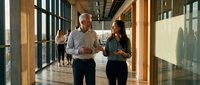 Professional middle-aged Caucasian businessman and young Asian businesswoman walking together in modern office corridor discussing work matters.